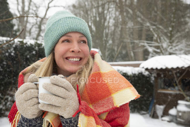 Весела жінка тримає чашку кави, яка дивиться далеко під час снігу — стокове фото