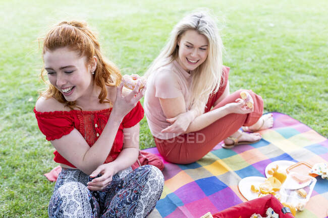 Усміхаючись, дівчата народжують пончик, сидячи на пікніковому покритті. — стокове фото
