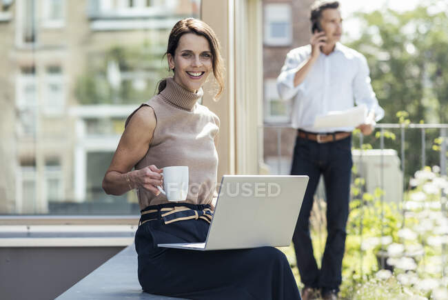 Lächelnde Geschäftsfrau hält Kaffeetasse in der Hand, während sie mit Kollegin im Hintergrund sitzt — Stockfoto