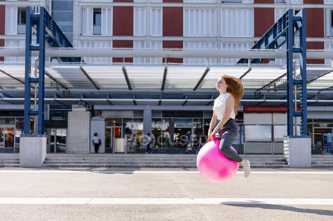 Carefree female freelancer jumping with bouncy ball by building during sunny day — Stock Photo