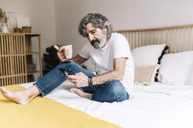 Uomo utilizzando smart phone in possesso di tazza di caffè mentre seduto sul letto a casa — Foto stock