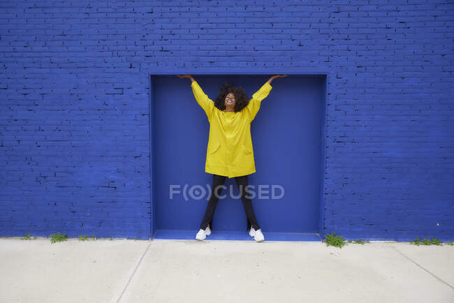 Mujer afro feliz de pie con los brazos levantados frente a la pared azul - foto de stock