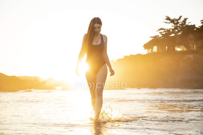 Modelo feminina em maiô preto andando na praia durante o pôr do sol — Fotografia de Stock