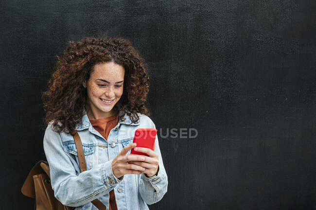 Lächelnde gelockte Frau mit Smartphone vor schwarzer Wand — Stockfoto