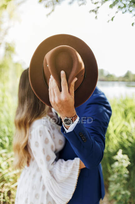 Man embracing girlfriend while hiding behind hat at park — Stock Photo