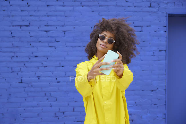 Beautiful young woman wearing eyeglasses taking selfie through mobile phone in front of blue wall — Stock Photo