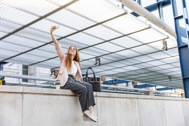Mujer de negocios sonriente tomando selfie a través del teléfono móvil mientras se sienta en el muro de contención en la estación de tren - foto de stock