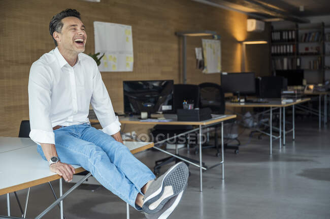 Feliz hombre de negocios sentado con las piernas cruzadas en el tobillo en el escritorio - foto de stock