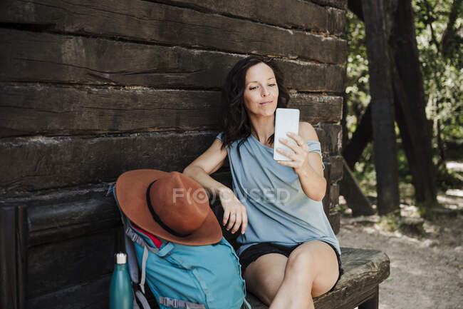 Взрослая женщина делает селфи через смартфон, сидя перед коттеджем в лесу — стоковое фото