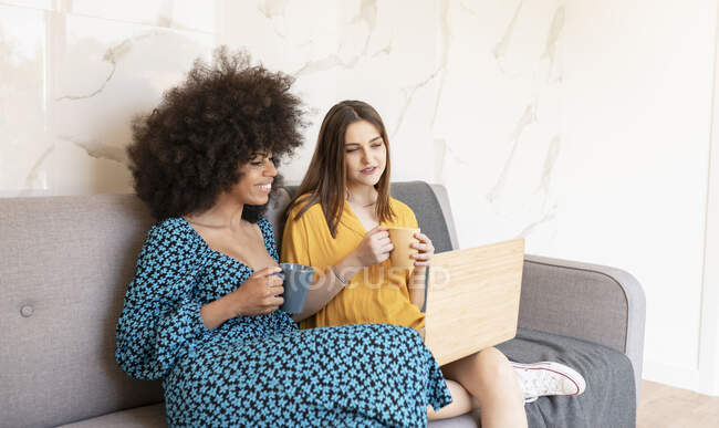 Young female friends looking at laptop while sitting on sofa at home — Stock Photo