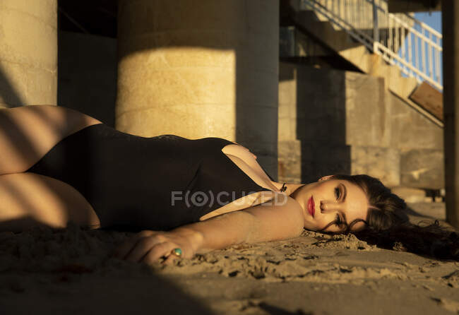 Modelo de moda sorridente em um maiô peça deitado na areia durante o pôr do sol — Fotografia de Stock
