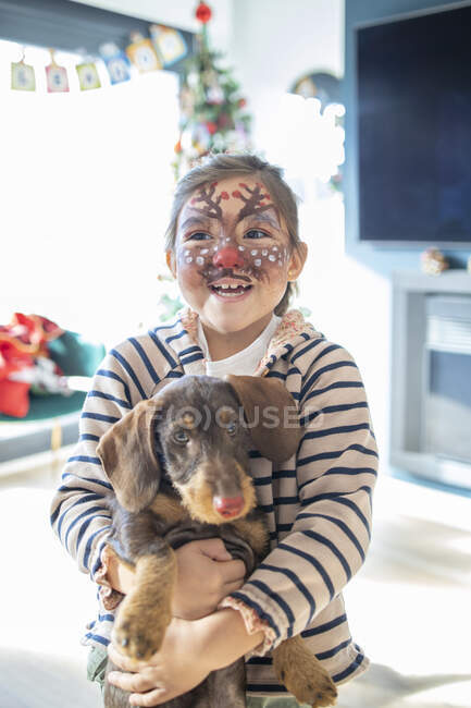 Улыбающаяся девушка с раскрашенным лицом держит собаку дома во время Рождества — стоковое фото