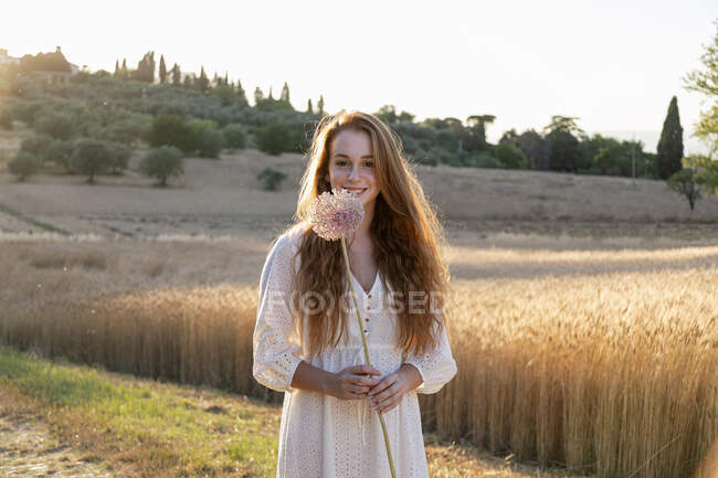 Mujer pelirroja sonriente sosteniendo flor de ajo en el campo - foto de stock