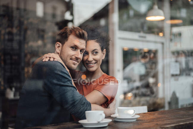 Pareja sonriendo mientras se abrazan mientras están sentados en la cafetería - foto de stock