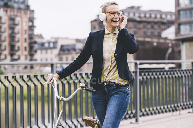 Счастливая профессиональная женщина слушает музыку через наушники, катаясь на велосипеде по мосту — стоковое фото