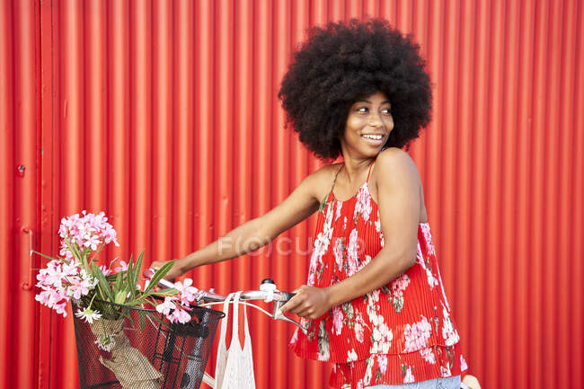 Afro donna con bicicletta sorridente mentre in piedi vicino al muro rosso — Foto stock
