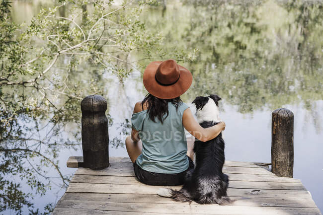 Frau mit Hut sitzt mit Arm um Hund auf Seebrücke — Stockfoto