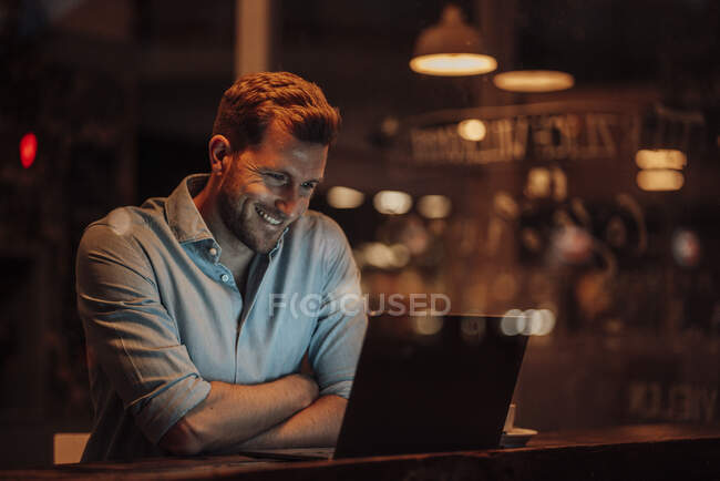 Hombre de negocios sonriente usando el ordenador portátil mientras está sentado en la cafetería - foto de stock