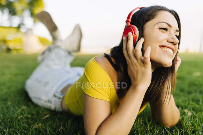 Mujer sonriente escuchando música a través de auriculares mientras se relaja en la hierba - foto de stock