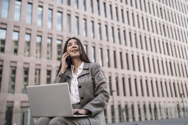 Mulher de negócios atenciosa falando no telefone inteligente ao usar o laptop contra a construção na cidade — Fotografia de Stock
