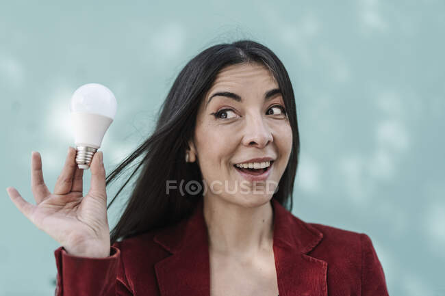 Empresária surpreendida segurando lâmpada contra parede azul-turquesa — Fotografia de Stock
