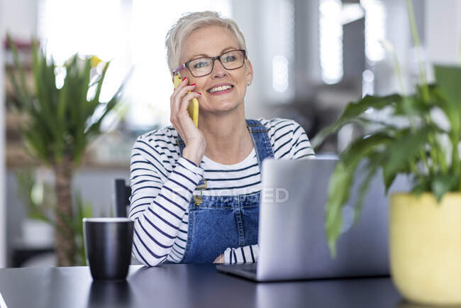 Mulher de negócios madura sorrindo enquanto fala no telefone inteligente no escritório em casa — Fotografia de Stock