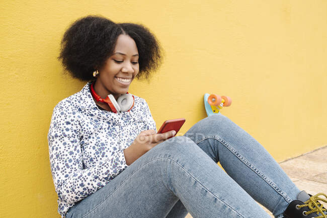 Mulher sorrindo mensagens de texto através do telefone celular no caminho da frente contra a parede — Fotografia de Stock