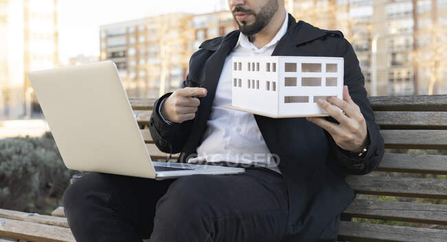 Чоловік архітектор тримає архітектурну модель під час відео дзвінка через ноутбук, сидячи на лавці в місті. — стокове фото