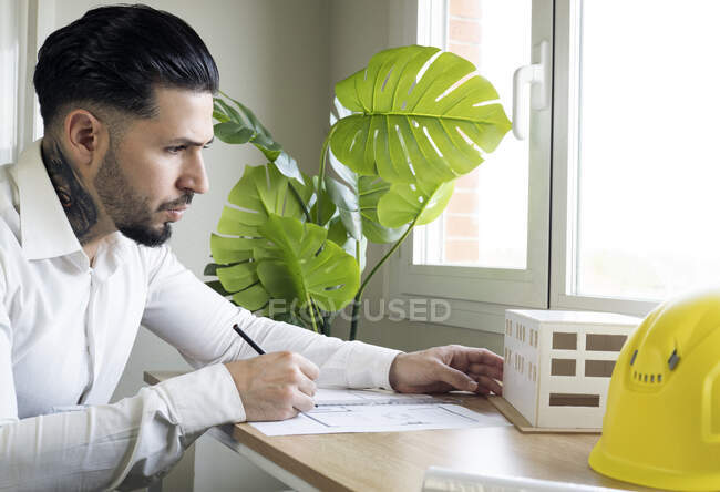 Männlicher Architekt schaut sich Architekturmodell an, während er im Büro eine Blaupause zeichnet — Stockfoto