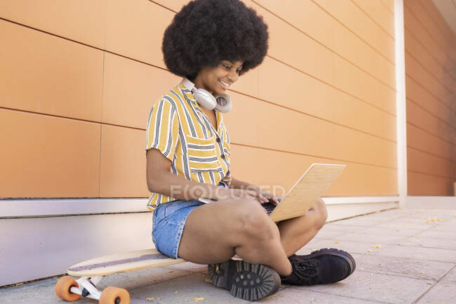Afro mujer usando el ordenador portátil mientras está sentado con las piernas cruzadas en el monopatín - foto de stock