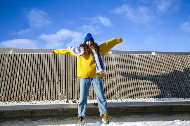 Chica sonriente con los brazos extendidos de pie contra la estructura durante el invierno - foto de stock