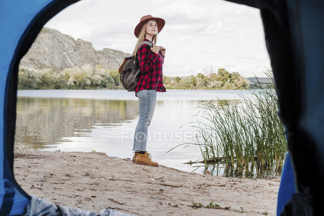 Giovane donna che indossa zaino guardando lontano in piedi sulla riva del lago — Foto stock