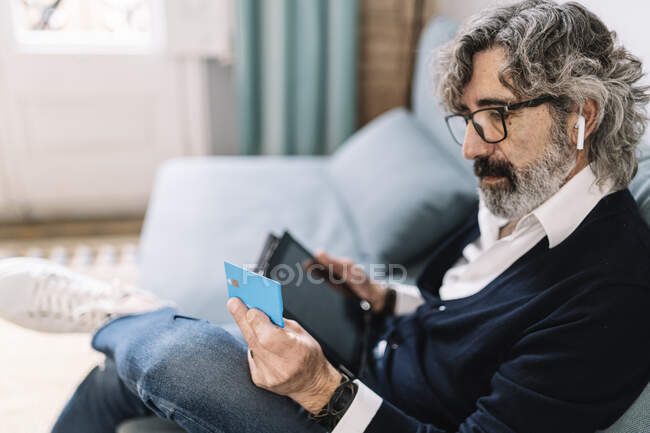 Красивый мужчина смотрит на кредитную карту с цифровым планшетом, сидя дома — стоковое фото