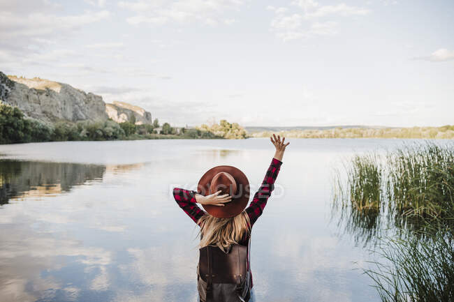 Giovane donna bionda che tiene il cappello in piedi con la mano sollevata sulla riva del lago — Foto stock