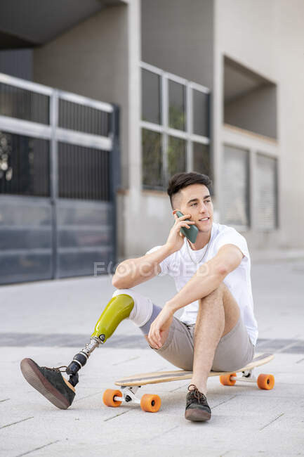 Jeune homme handicapé parlant par téléphone portable tout en étant assis sur le skateboard — Photo de stock