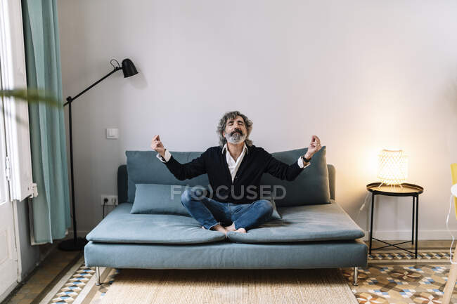 Старший медитирует, сидя дома на диване — стоковое фото