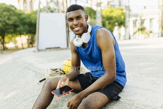 Smiling man holding smart phone while sitting at basketball court — Stock Photo