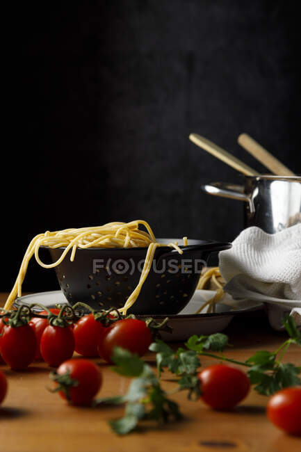 Spaghetti in colander amidst fresh cherry tomatoes and cilantro on wooden table — Stock Photo