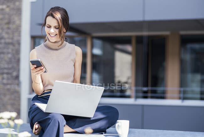 Businesswoman with laptop using mobile phone while sitting on retaining wall — Stock Photo