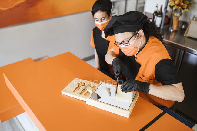 Waitress looking at female chef cutting cheese on board during COVID-19 — Stock Photo