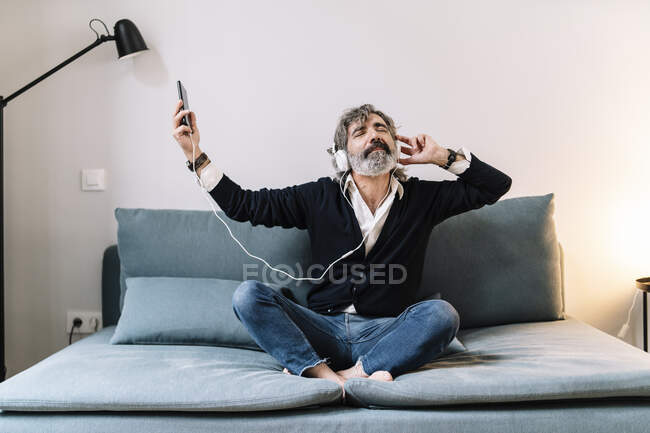 Uomo anziano che ascolta musica attraverso le cuffie mentre è seduto sul divano a casa — Foto stock