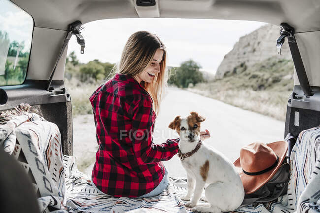 Happy blond woman sitting with dog in car trunk — Stock Photo