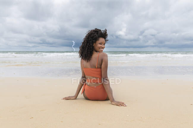 Sorridente giovane donna guardando indietro sopra la spalla mentre seduto in spiaggia — Foto stock