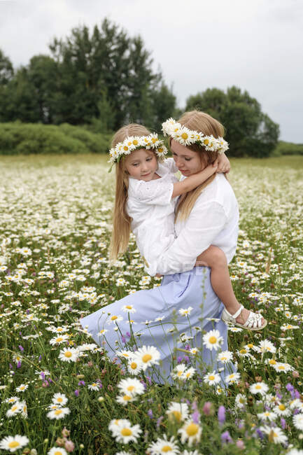 Woman with flower tiaras carrying daughter while walking at chamomile field — Stock Photo