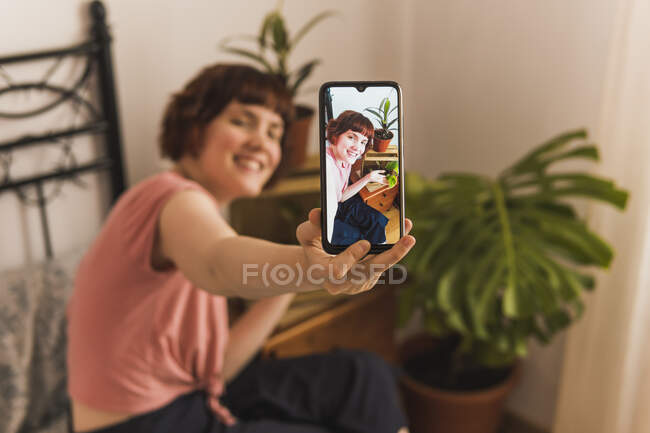 Donna sorridente che prende selfie attraverso il telefono cellulare seduto in camera da letto a casa — Foto stock