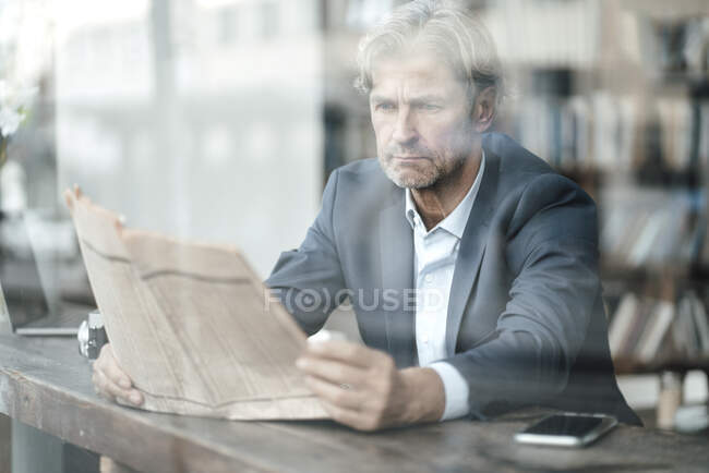 Maduro hombre de negocios leyendo periódico sentado en la cafetería visto a través de la ventana de vidrio - foto de stock