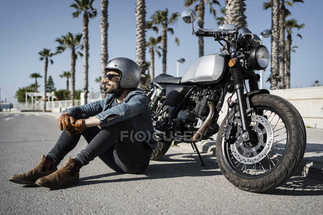 Jeune homme portant un casque assis devant la moto sur la route — Photo de stock
