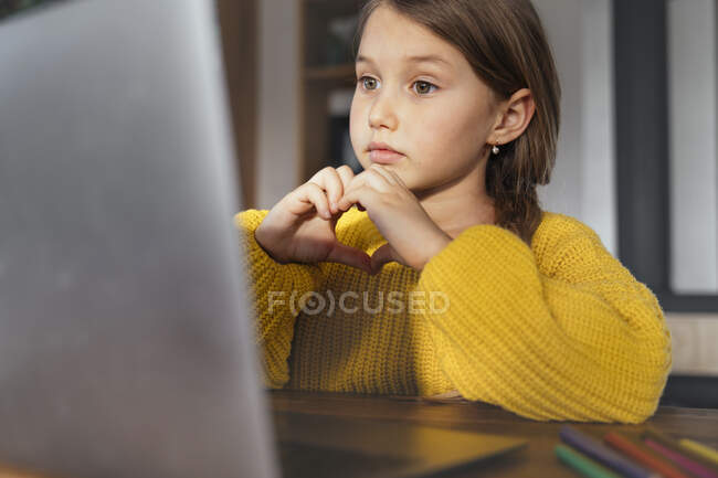 Милая маленькая девочка, придает форму сердцу рукой во время видеозвонка на ноутбук дома — стоковое фото