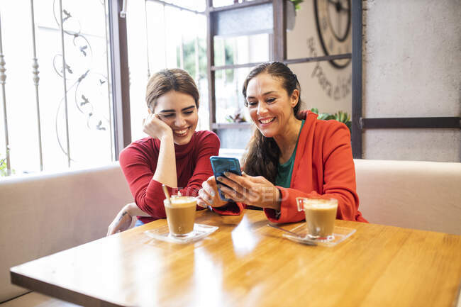Mujer alegre mostrando el teléfono móvil a una amiga en la cafetería - foto de stock