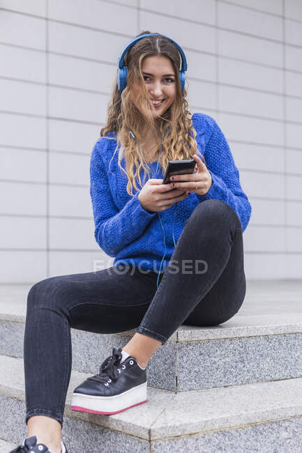 Mujer sonriente escuchando música a través de auriculares mientras está sentada en los escalones - foto de stock